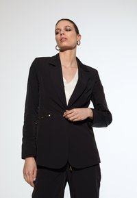 Black blazer with a fitted design, featuring gold-tone accent buttons and a notched collar. Worn over a cream top. Smooth fabric texture.