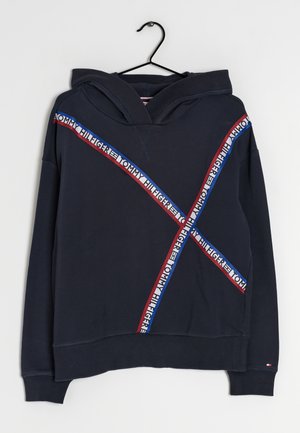 Granatowa bluza z kapturem z przeplatanymi czerwonymi, białymi i niebieskimi taśmami z logo Tommy Hilfiger na przodzie i rękawach, wisząca na czarnym wieszaku.