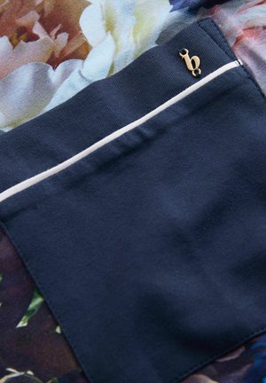 Marineblauer Stoff mit floralem Muster, ausgestattet mit einer kleinen Tasche mit weißer Umrandung und goldenem Akzent. Durchgehend glatte Textur.