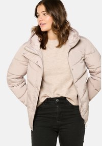 Doudoune rose pâle avec fermeture éclair à l'avant, capuche et fermetures à pression. Présente un design matelassé et une texture douce et lisse. Portée sur un pull beige.