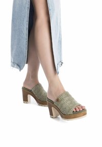 Mule verdi intrecciati con tacco e punte aperte, caratterizzati da una piattaforma, suola in gomma beige e un piccolo bottone decorativo sul lato.