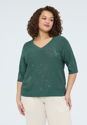 Žena s kudrnatými vlasy, oblečená v tmavě zeleném svetru s výstřihem do V a malými bílými skvrnami, a světlých kalhotách, stojící před jednolitým pozadím.