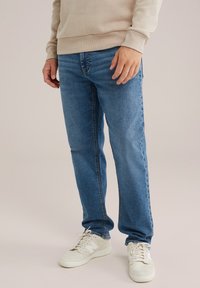 Zilā džinsu bikses ar taisnu piegriezumu, viegli izbalējušas virsmas un tonāliem šuvēm, kombinētas ar bežām kedām un bežīgu sporta krekls.