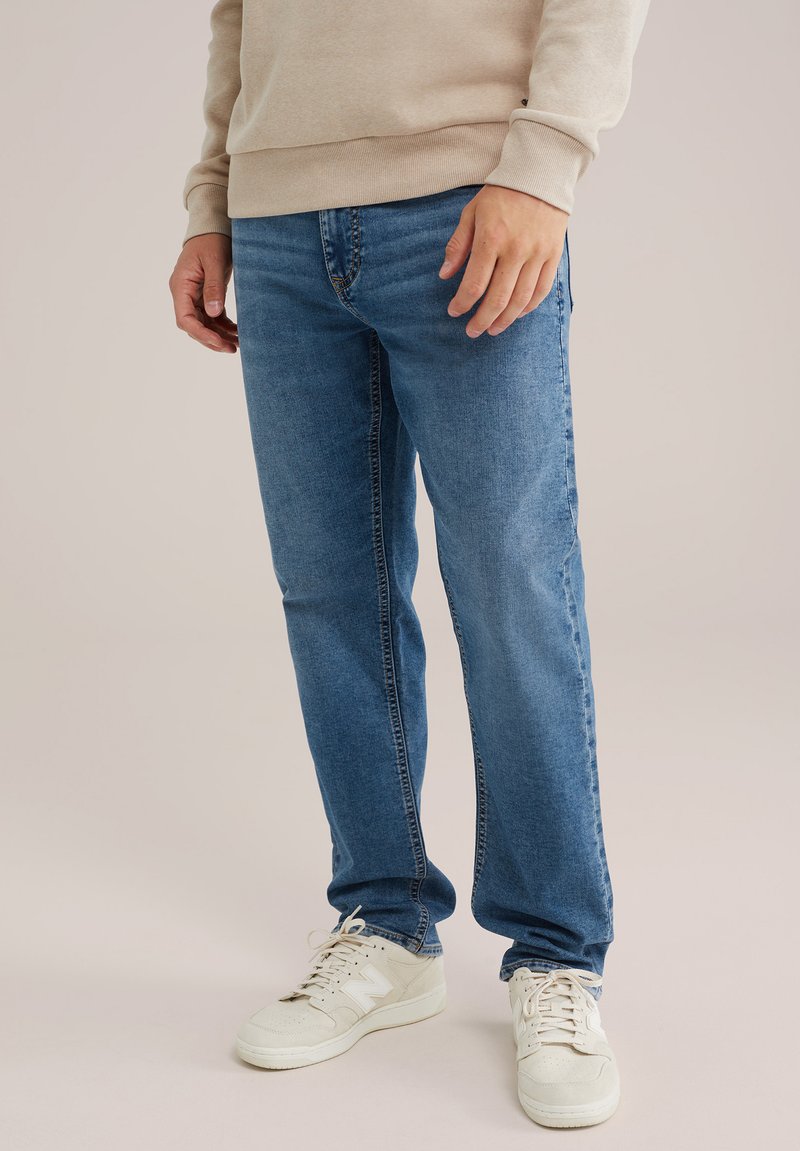 Zilā džinsu bikses ar taisnu piegriezumu, viegli izbalējušas virsmas un tonāliem šuvēm, kombinētas ar bežām kedām un bežīgu sporta krekls.