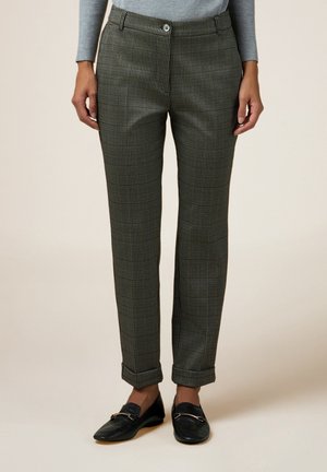 Frau trägt graue, gemusterte, maßgeschneiderte Hose mit umgeschlagenen Säumen und schwarzen Slippern, steht vor beigem Hintergrund.