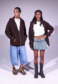 Felpa marrone zip-up, t-shirt bianca, ampi pantaloni di jeans blu; abbinati a una gonna di jeans e stivali neri alti fino al ginocchio. Look casual da strada.