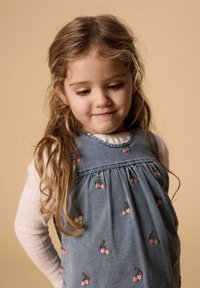 Nuori tyttö, jolla on pitkät vaaleat hiukset, pukeutunut farkkumekkoon, jossa on kirsikkaompeleita, ja valkoiseen pitkähihainen puseroon, katsoo alas kevyesti hymyillen.