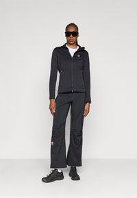 Chaqueta negra con cremallera y dos bolsillos delanteros, con un logo; combinada con pantalones negros de corte holgado que presentan un parche de logo rojo. Zapatillas negras y gafas de sol.