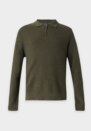 Uomo in piedi che indossa un maglione verde oliva lavorato a maglia, pantaloni a quadri marroni, scarpe da ginnastica bianche e una camicia a righe sotto al maglione.