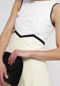 Valkoinen ja keltainen mekko mustilla yksityiskohdilla, jossa on vartalonmyötäinen yläosa ja levenevä hameosa. Musta clutch-laukku pidetään oikeassa kädessä.