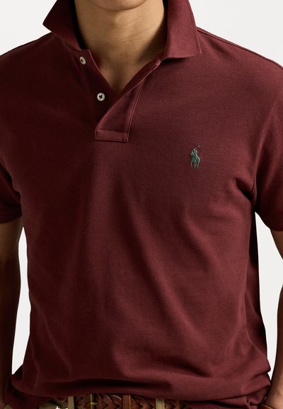 Polo de burgundă din țesătură texturată, având o deschidere cu trei butoane și un logo brodat verde pe pieptul stâng.