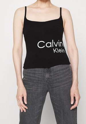 Kobieta ubrana w czarny top z napisem „Calvin Klein” z przodu oraz wyblakłe czarne dżinsy, stojąca na tle prostego białego tła.