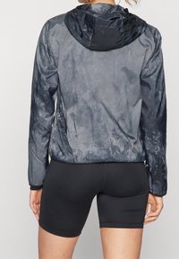 Giacca leggera con cappuccio in tessuto grigio scuro, dalla trama texture con una finitura leggermente lucida. Presenta tasche con cerniera e polsini elasticizzati; progettata per un abbigliamento attivo.