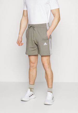 Mann trägt beige Adidas-Shorts mit weißen Streifen, weiße Adidas-Sneakers, passende beige gestreifte Socken und ein weißes T-Shirt.