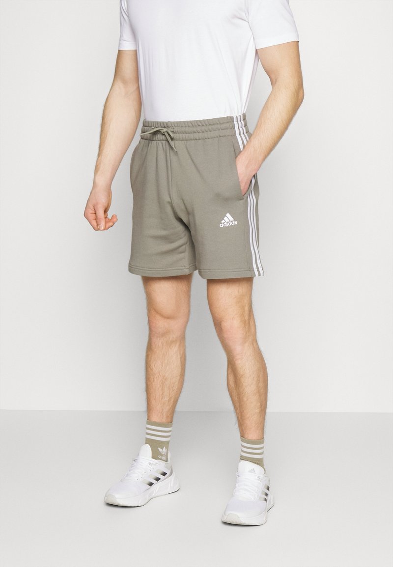 Mann trägt beige Adidas-Shorts mit weißen Streifen, weiße Adidas-Sneakers, passende beige gestreifte Socken und ein weißes T-Shirt.