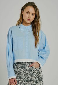 Camicia azzurro chiaro a maniche lunghe, cropped, con chiusura a bottoni sul davanti, taschino singolo sul petto e dettaglio bianco sull'orlo. Abbinata a pantaloni a stampa serpente texturizzati.