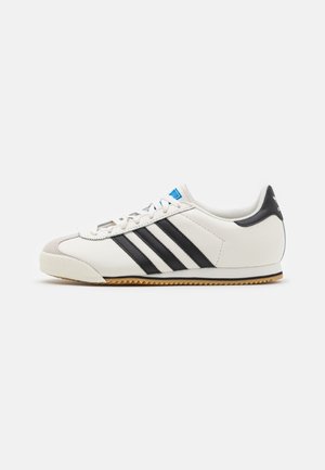 Baskets basses blanches avec bandes noires, semelle en gomme et lacets blancs, vues de côté sur un fond clair uni.
