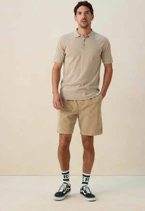 Beige poloshirt met korte mouwen, khaki shorts en zwarte sneakers met witte accenten. Sokken met groene tekst "RER" zichtbaar boven de sneakers.