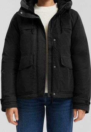 Personne portant une veste d'hiver noire à capuche sur un pull blanc et un jean bleu, debout devant un fond uni.