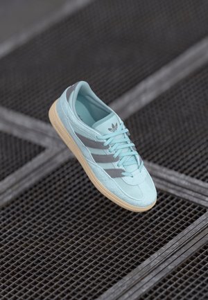 Baskets Adidas bleu clair avec des rayures grises et une semelle beige, posées au-dessus d'une surface en grille métallique noire.