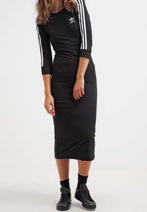 Kobieta ubrana w czarną sukienkę midi Adidas z rękawami 3/4 i białymi paskami, w czarnych skarpetkach i czarnych tenisówkach, stojąca na białej podłodze.
