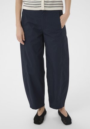 Osoba ubrana w granatowe spodnie o szerokich nogawkach, czarne siateczkowe buty wsuwane oraz biały, bezrękawnik w czarne paski, stojąca z jedną ręką w kieszeni.