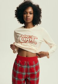 Kremowa bluzka z długim rękawem z grafiką "Stay Cool" w kolorze czerwonym, w połączeniu z czerwoną piżamą w kratę z zielonymi i białymi liniami. Miękki materiał, luźny krój.