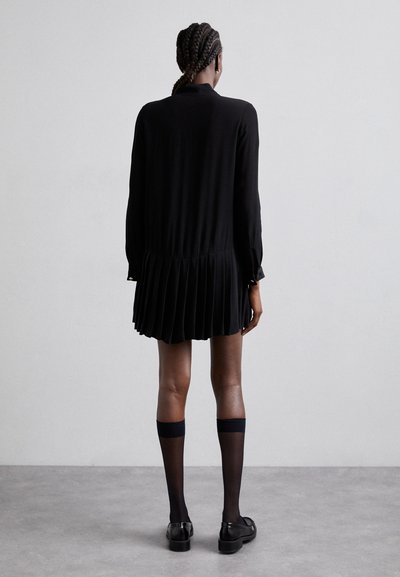 Robe noire plissée avec manches longues et col. Tissu lisse, longueur genoux, associée à des chaussettes noires jusqu'aux genoux et des chaussures noires.