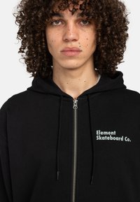 Black zip-up hoodie with drawstrings, featuring a light green logo "Element Skateboard Co." on the left chest. Soft fabric, casual design.