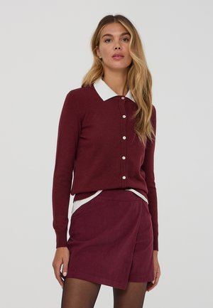 Cardigan bordeaux con colletto bianco, con bottoni sulla parte anteriore. Abbinato a una gonna in velluto a coste dello stesso colore, entrambi in tonalità simili.