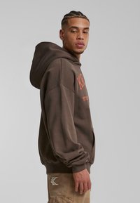Sudadera de polar marrón con un bolsillo frontal, logo en naranja y un corte relajado. Cuenta con una gran capucha ajustable y puños acanalados.