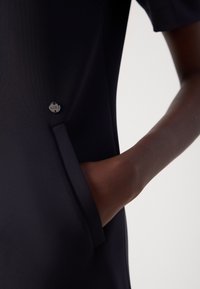 Robe noire avec une poche ornée d'un bouton en argent. Le matériau semble lisse, avec une forme structurée et des manches courtes.