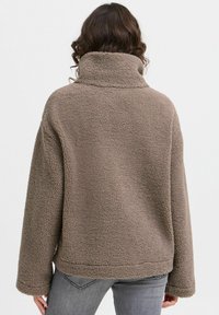 Brun fleecejakke med høj krave, afslappet pasform og lange ærmer. Textureret ydre og ribbede manchetter med rundet kant.