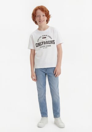 Niño pelirrojo sonriente con una camiseta blanca con diseño gráfico, vaqueros azul claro y zapatillas blancas, de pie frente a un fondo blanco.