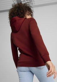 Felpa bordeaux con cappuccio foderato, polsini e orlo a costine. Il modello indossa jeans azzurri chiari. La texture appare liscia; design minimalista su uno sfondo semplice.