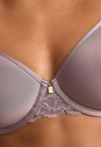 Violetinė liemenėlė su lygiu audiniu, nėrinių apdaila apačioje ir mažu auksiniu ženkleliu su raide „N“ viduryje priekinėje dalyje.