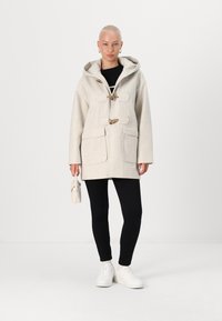 Beige ullfrakk med hette, snørelukking og to frontlommer. Båret over svarte leggings og hvite sneakers, holdt med en liten veske.