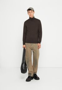 Bruine coltrui, lichtbeige denim broek, zwarte chunky schoenen en een grijze pet. Houdt een zwarte leren tas vast. Minimalistische achtergrond.