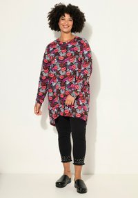 Tunika a maniche lunghe con un motivo di labbra colorate e testo, abbinata a pantaloni neri a 3/4 con dettagli borchiati e zoccoli neri.