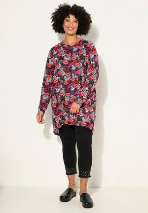 Túnica de manga larga con un patrón de labios coloridos y texto, combinada con pantalones negros recortados con acentos de tachuelas y zuecos negros.