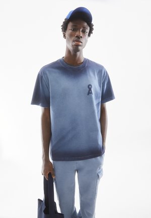 Blauwe gradient t-shirt met een cirkelvormig logo; gedragen met lichtblauwe broek. Model houdt een bijpassende donkerblauwe totebag vast, tegen een witte achtergrond.