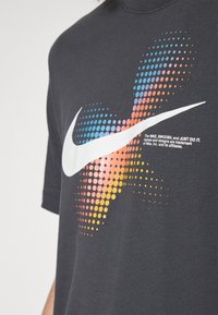 Šedé bavlněné tričko s velkým bílým logem Nike Swoosh nad tečkovaným gradientním vzorem v modré, oranžové a žluté barvě. Text obsahuje podrobnosti o ochranné známce.