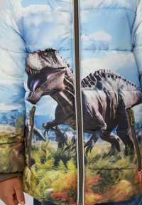 Flauschiger blauer Mantel mit einem Dinosaurier-Print auf der Vorderseite, strukturiertem Oberflächenmaterial, Reißverschluss und kontrastierenden braunen Reißverschlussakzenten.