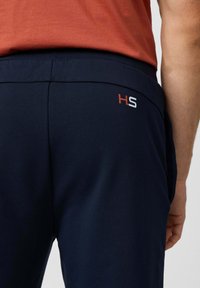 Donkerblauwe sportbroek met een gladde textuur, voorzien van een rood en wit "HS"-logo op de tailleband. Het ontwerp heeft een comfortabele pasvorm.