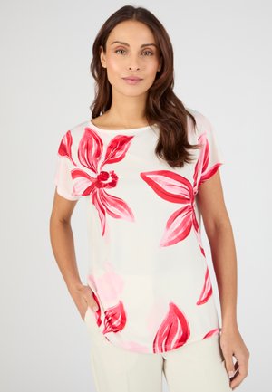 Donna con lunghi capelli castani che indossa una camicetta bianca a maniche corte con grandi stampe floreali rosse, in piedi con una mano in tasca.