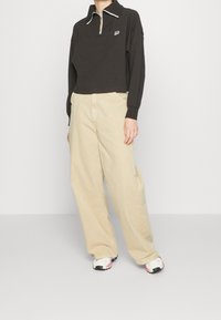 Pullover corto marrone scuro con colletto bianco e chiusura con zip, abbinato a pantaloni beige a gamba larga e sneaker con motivo.
