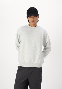 Felpa grigia chiara con colletto rotondo, polsini e orlo a costine; indossata con pantaloni cargo neri e un cappellino da baseball nero.