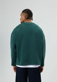Donkergroene oversized trui met ronde hals en verzakte schouders, gestructureerde stof, gedragen over een wit shirt, gecombineerd met donkere spijkerbroek.