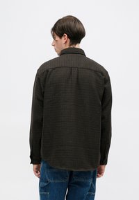 Cămașă maro cu model, cu guler cu nasturi, mâneci lungi și tiv tăiat. Pur tată cu pantaloni cargo albaștri, care arată textura în carouri.