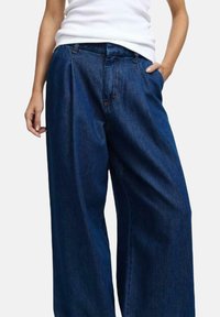 Pantaloni in denim blu a gamba larga con vita alta, caratterizzati da pieghe frontali, zip e tasche. Texture morbida, leggermente usurata.